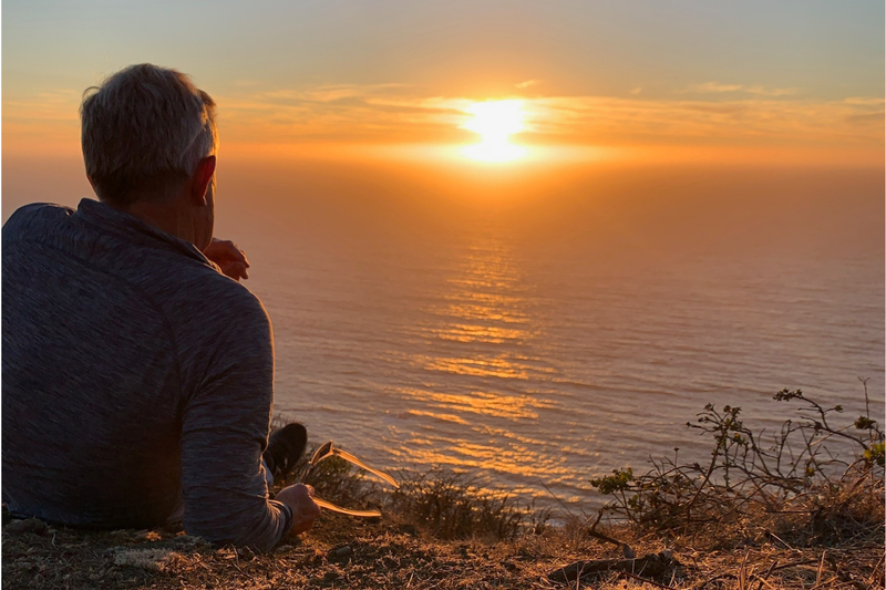 Man sitting on a coastal cliff at sunset overlooking the ocean, reflecting Lighthouse Lane Books’ themes of journey and discovery.