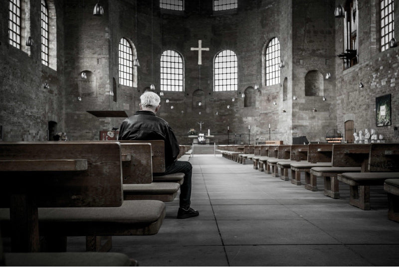 A lone man sitting on a wooden pew in an empty, dimly lit stone church, reflecting on grief, faith, and the closures of organized religion facilities like the Oakland Diocese.
